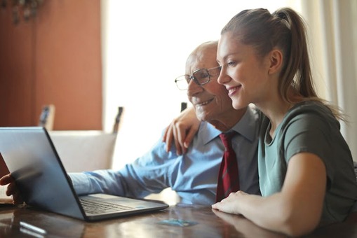 A young, positive woman helps an older man using a laptop.